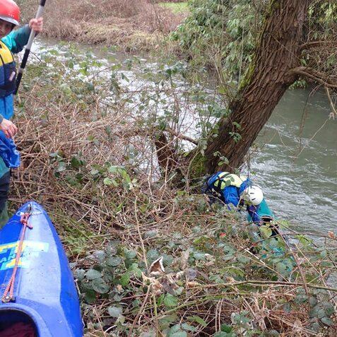Zwei Kajakfahrer am Ufer eines Flusses, einer klettert zwischen Str&auml;uchern, ein Kajak liegt im Vordergrund.