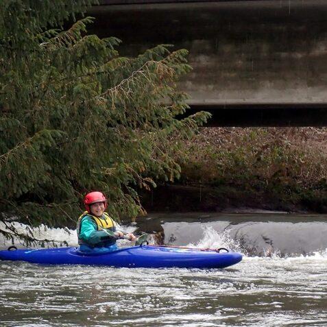 Kajakfahrer in blauer Kajak mit rotem Helm paddelt auf einem schmalen Fluss unter einer Br&uuml;cke.