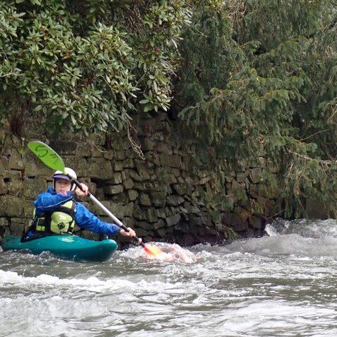 Kajakfahrer paddelt auf einem Gew&auml;sser neben einer steinernen Uferlinie, umgeben von &uuml;ppigem Gr&uuml;n.