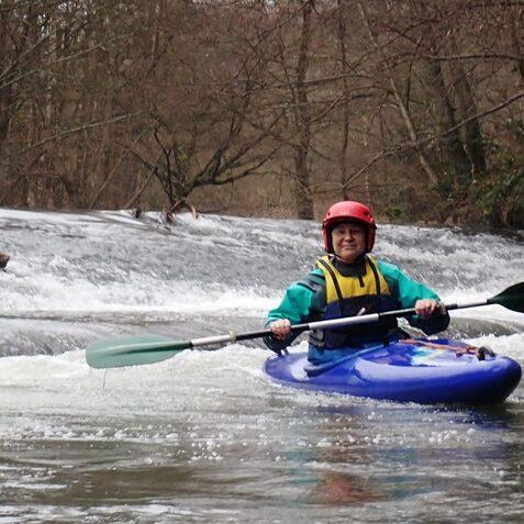 Junge f&auml;hrt mit einem blauen Kajak auf einem ruhigen Fluss, umgeben von B&auml;umen und einer kleinen Wasserfallstelle.