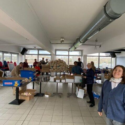 Gruppensituation in einem Raum mit Tischen voller Kartons und Leuten, die verschiedene Gegenst&auml;nde organisieren.