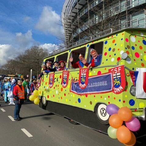 Bunt gestalteter VW-Bus mit feiernden Menschen auf einer Karnevalsparade, umgeben von Teilnehmern in bunter Kleidung.