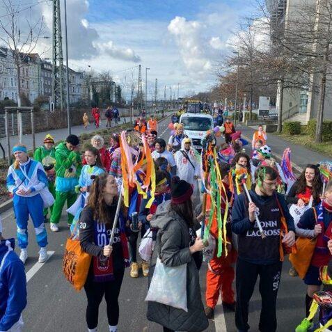 Bunte Menschen in Kost&uuml;men stehen auf einer Stra&szlig;e und feiern, umgeben von festlicher Dekoration und fr&ouml;hlicher Stimmung.