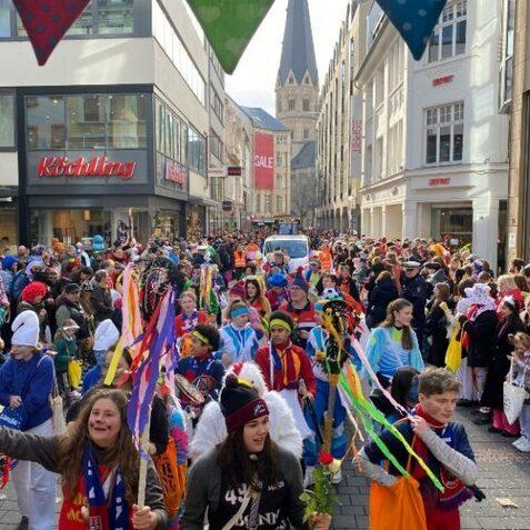 Bunte Menschenmenge in Kost&uuml;men mit bunten B&auml;ndern in einer Stadtstra&szlig;e, im Hintergrund eine Kirche und Gesch&auml;fte.