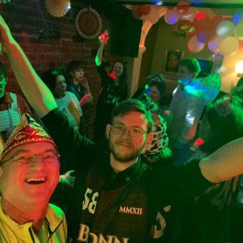 Gruppe von feiernden Menschen in einem bunten Raum, mit einer Person im Vordergrund, die l&auml;chelt und die Arme hebt.