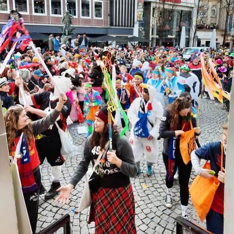 Bunte Karnevalsfeier mit verkleideten Menschen, die farbige F&auml;hnchen schwenken und in einem festlichen Treiben sind.