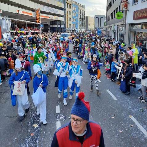 Karnevalsumzug mit kost&uuml;mierten Menschen, die fr&ouml;hlich durch eine belebte Stra&szlig;e ziehen, umgeben von Zuschauern.