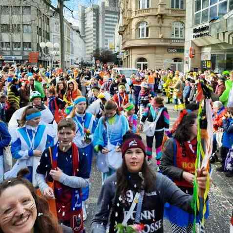 Bunte Menschenmenge feiert Karneval auf einer Stra&szlig;e, mit Konfetti und festlicher Kleidung. St&auml;dtereferenz im Hintergrund.