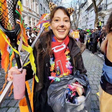 Frau mit Tierohren und bunter Dekoration h&auml;lt ein Getr&auml;nk und eine T&uuml;te voller Kost&uuml;mschmuck auf einem Fest.