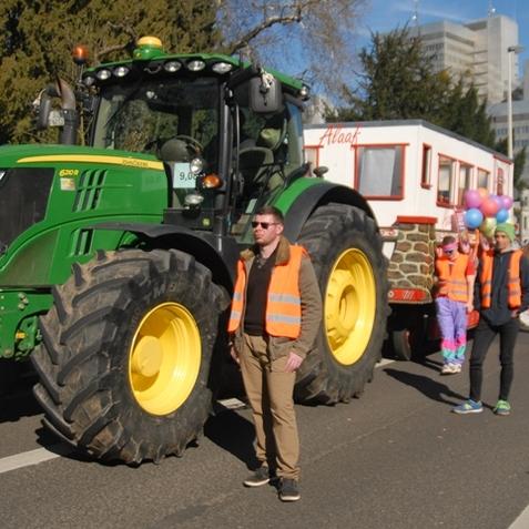 Traktor mit gro&szlig;em Reifensatz und bunten Festwagen bei einer Stra&szlig;enparade, umgeben von verkleideten Teilnehmern.