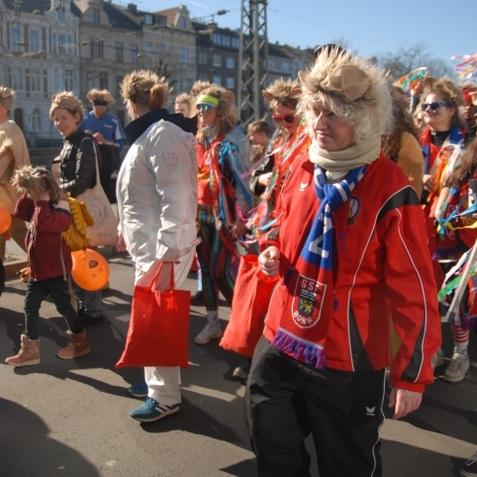 Menschen in bunten Kost&uuml;men und Per&uuml;cken ziehen in einer festlichen Parade mit Taschen und B&auml;ndern auf einer Stra&szlig;e entlang.