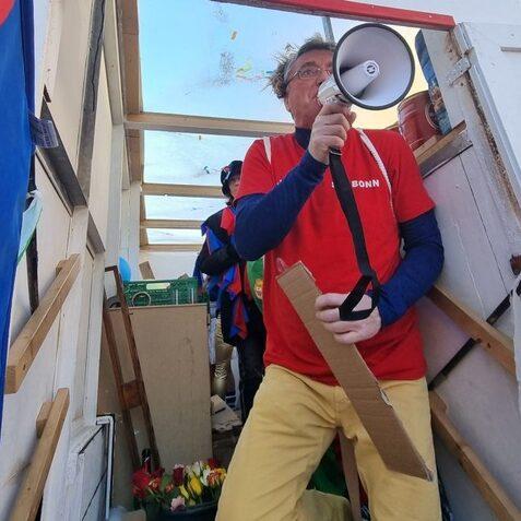 Mann in rotem T-Shirt h&auml;lt ein Megafon, w&auml;hrend er im Hintergrund von bunten Kost&uuml;men umgeben ist.