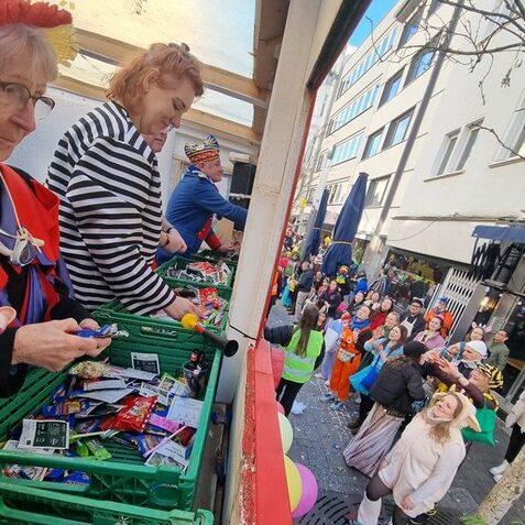 Blick auf eine Karnevalsfeier: Menschen werfen S&uuml;&szlig;igkeiten vom Wagen in eine jubelnde Menge auf der Stra&szlig;e.