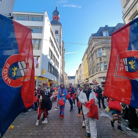 Karnevalsumzug in Bonn mit Menschen in Kost&uuml;men und Fahnen in einer belebten Stra&szlig;e. Im Hintergrund ist ein Kirchturm sichtbar.