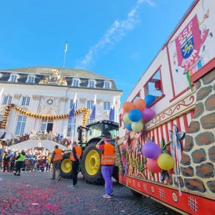 Bunte Festwagen und einen Traktor auf einer Stra&szlig;e vor einem festlich dekorierten Geb&auml;ude w&auml;hrend einer Parade.