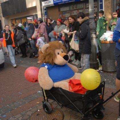 Gro&szlig;er Pl&uuml;schl&ouml;we in blauer Kleidung sitzt in einem Kinderwagen, umgeben von feiernden Menschen und bunten Luftballons.