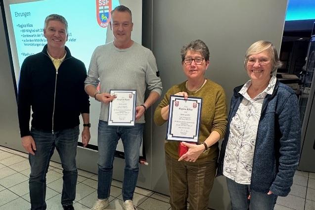 Vier Personen stehen in einem Raum und halten Zertifikate in den H&auml;nden, im Hintergrund ein Projektorbild.