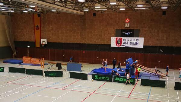 Sporthalle mit Ger&auml;ten f&uuml;r Gymnastiktraining, in der mehrere Jugendliche aktiv sind. Ein Banner mit &bdquo;Mein Verein am Rhein&ldquo; ist sichtbar.