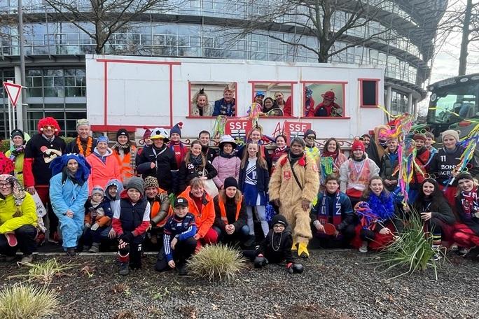 Gruppenselfie von Kost&uuml;mierten vor einem festlich geschm&uuml;ckten Wagen und einem Traktor, umgeben von B&auml;umen und Geb&auml;uden.