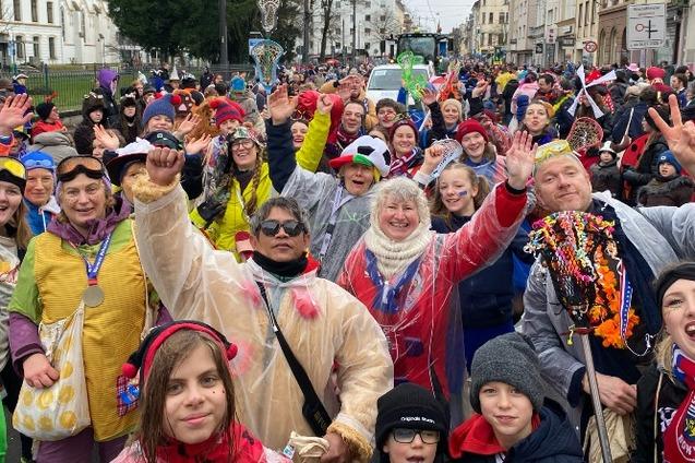 Gruppe von fr&ouml;hlichen Menschen in bunten Kost&uuml;men feiert auf der Stra&szlig;e, einige winken, andere l&auml;cheln in die Kamera.