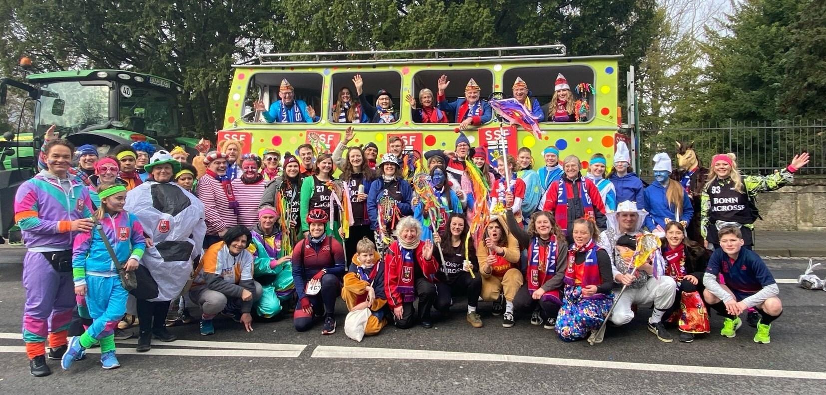 Gruppenselfie von Festteilnehmern in bunten Kost&uuml;men vor einem farbenfrohen Festivalbus.
