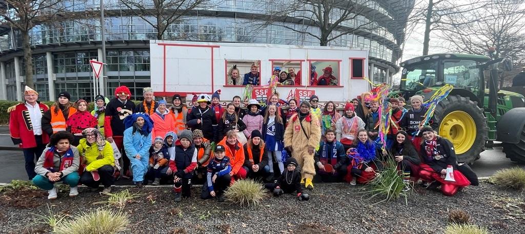 Gruppenselfie von Kost&uuml;mierten vor einem festlich geschm&uuml;ckten Wagen und einem Traktor, umgeben von B&auml;umen und Geb&auml;uden.