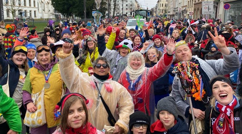 Gruppe von fr&ouml;hlichen Menschen in bunten Kost&uuml;men feiert auf der Stra&szlig;e, einige winken, andere l&auml;cheln in die Kamera.