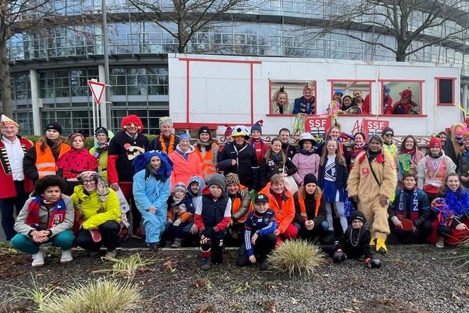 Gruppenselfie von Kost&uuml;mierten vor einem festlich geschm&uuml;ckten Wagen und einem Traktor, umgeben von B&auml;umen und Geb&auml;uden.