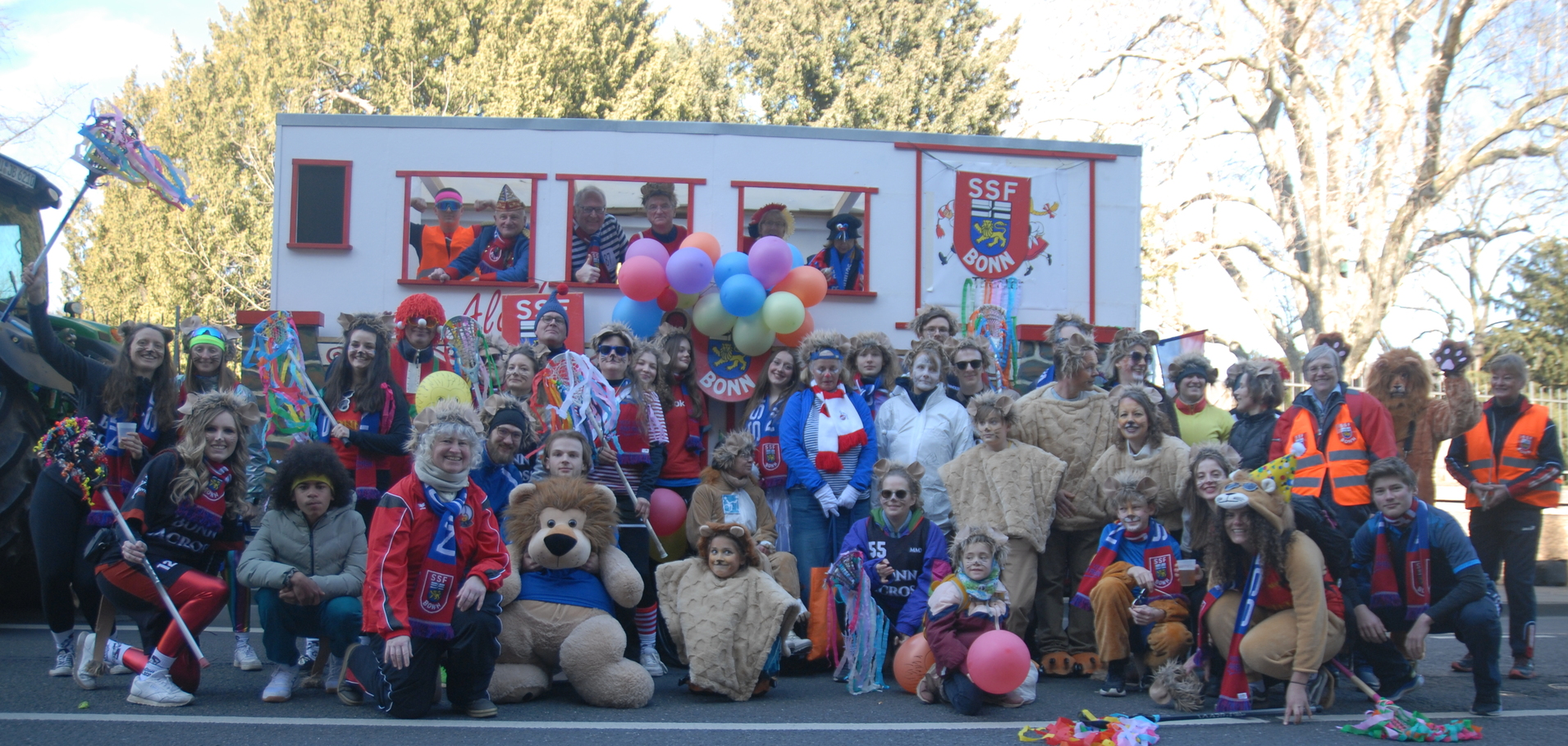 Gruppe von verkleideten Menschen vor einem bunten Festwagen mit Luftballons und Suits, in einer festlichen Umgebung.