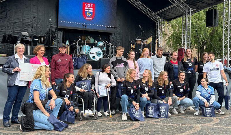 Gruppenszene mit Sportlerinnen des SSF Bonn auf einer B&uuml;hne, umgeben von Zuschauern und instrumenteller Band.
