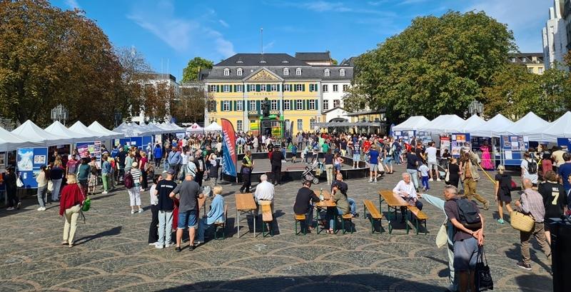Belebter Marktplatz mit Verkaufsst&auml;nden, Besuchern und historischer Architektur im Hintergrund.