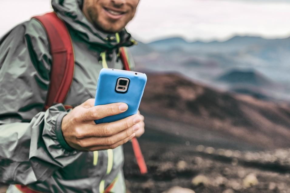 Mann in wetterfester Jacke und Rucksack h&auml;lt ein Smartphone vor einer vulkanischen Landschaft.