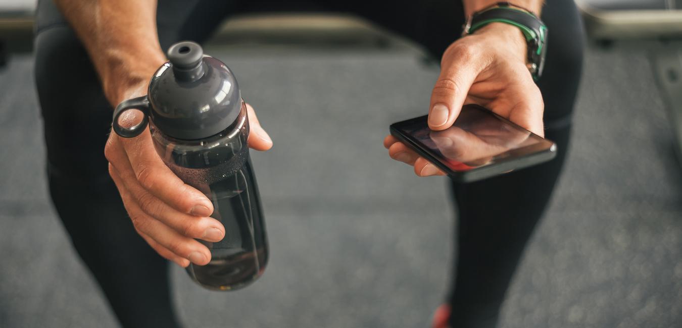 Nahaufnahme einer Hand mit einer Wasserflasche und eines Smartphones, sitzend auf einem Fitnessger&auml;t.