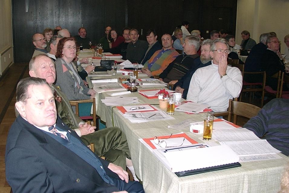 Gruppenszene bei einer Versammlung mit mehreren Personen an Tischen, einige trinken Bier, Papiere liegen verteilt.