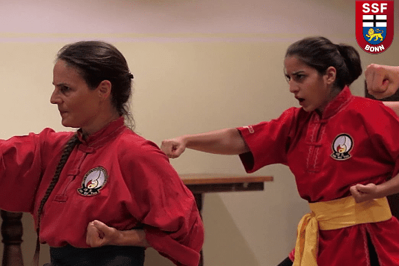 Zwei Kampfsportlerinnen in roten und schwarzen Anz&uuml;gen f&uuml;hren eine Technik&uuml;bung in einem Trainingsraum aus.