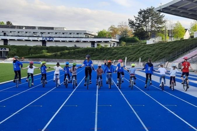 Gruppe von Kindern und Erwachsenen auf Einr&auml;dern, die auf blauer Rennbahn stehen und H&auml;nde halten.