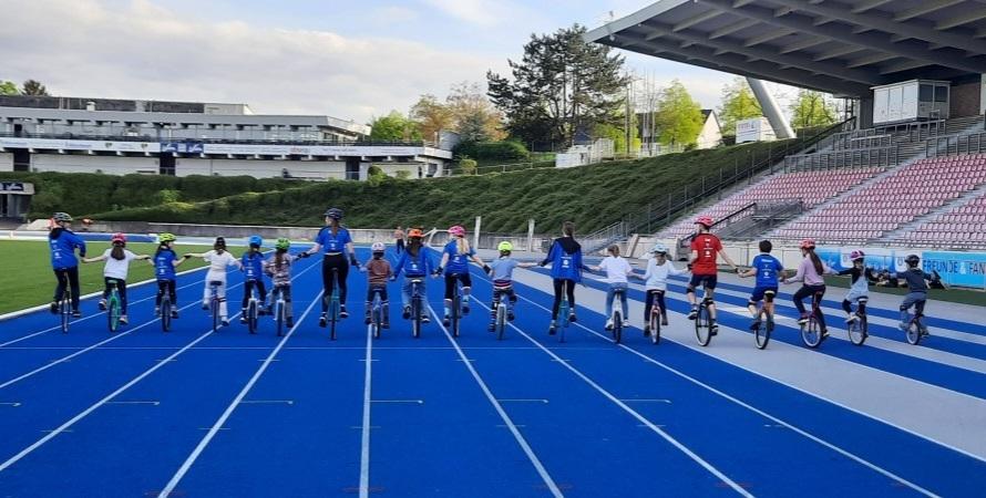 Gruppe von Kindern und Erwachsenen auf Einr&auml;dern, die auf blauer Rennbahn stehen und H&auml;nde halten.