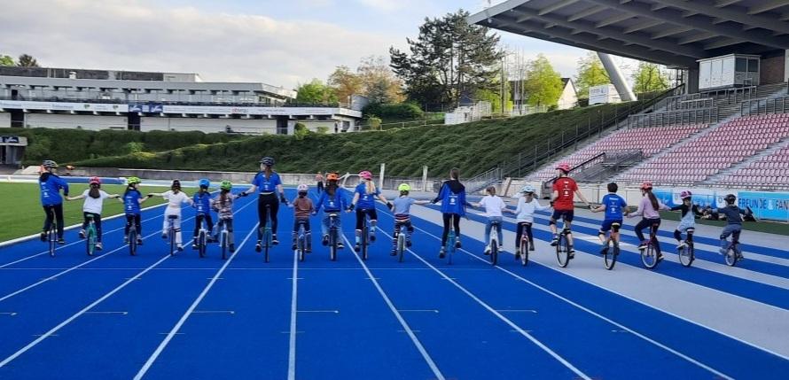 Gruppe von Kindern und Erwachsenen auf Einr&auml;dern, die auf blauer Rennbahn stehen und H&auml;nde halten.