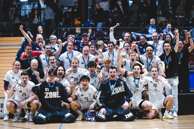 Gruppenszene mit jubelnden Handballspielern in Trikots, die stolz Medaillen tragen und den Sieg feiern.