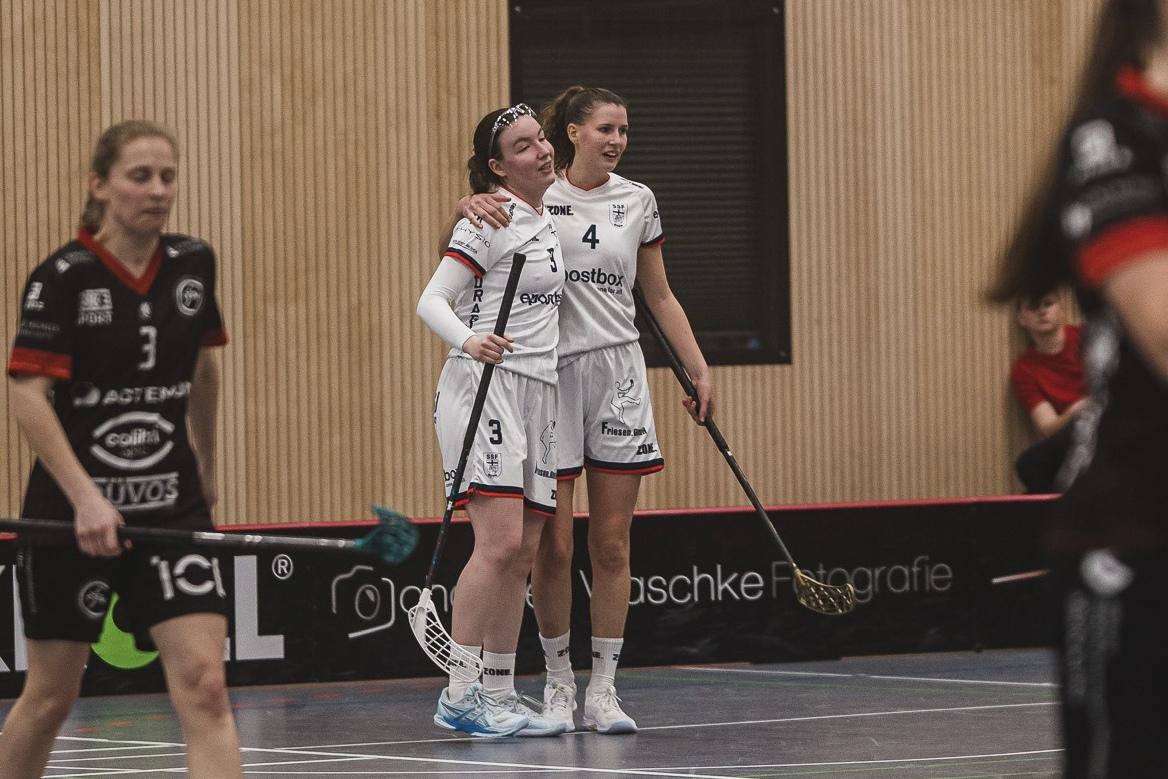 Zwei Spielerinnen im wei&szlig;en Trikot umarmen sich freudig auf einem Hallenboden w&auml;hrend eines Floorball-Spiels.
