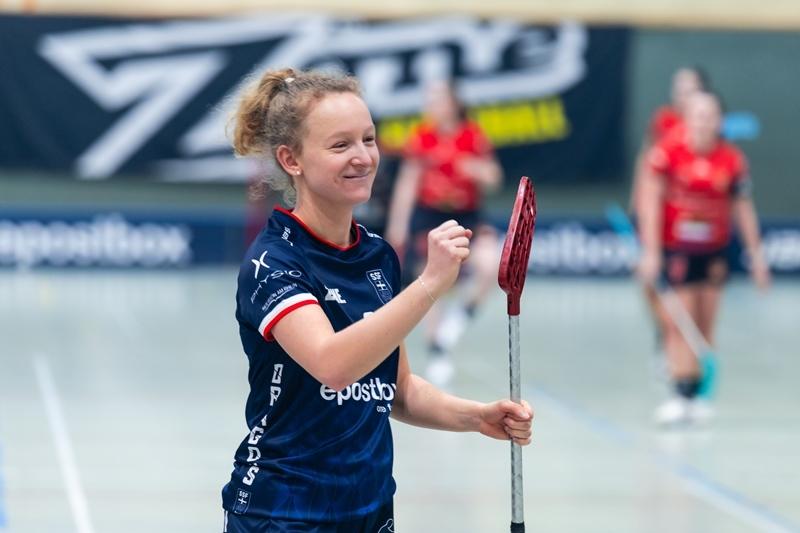 Freudige Spielerin mit Floorball-Schl&auml;ger in sportlicher Kleidung, Hintergrund zeigt weitere Spielerinnen in Aktion.