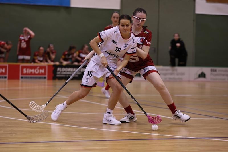 Zwei Spielerinnen im Floorball k&auml;mpfen um den Ball, w&auml;hrend Zuschauer im Hintergrund zuschauen.