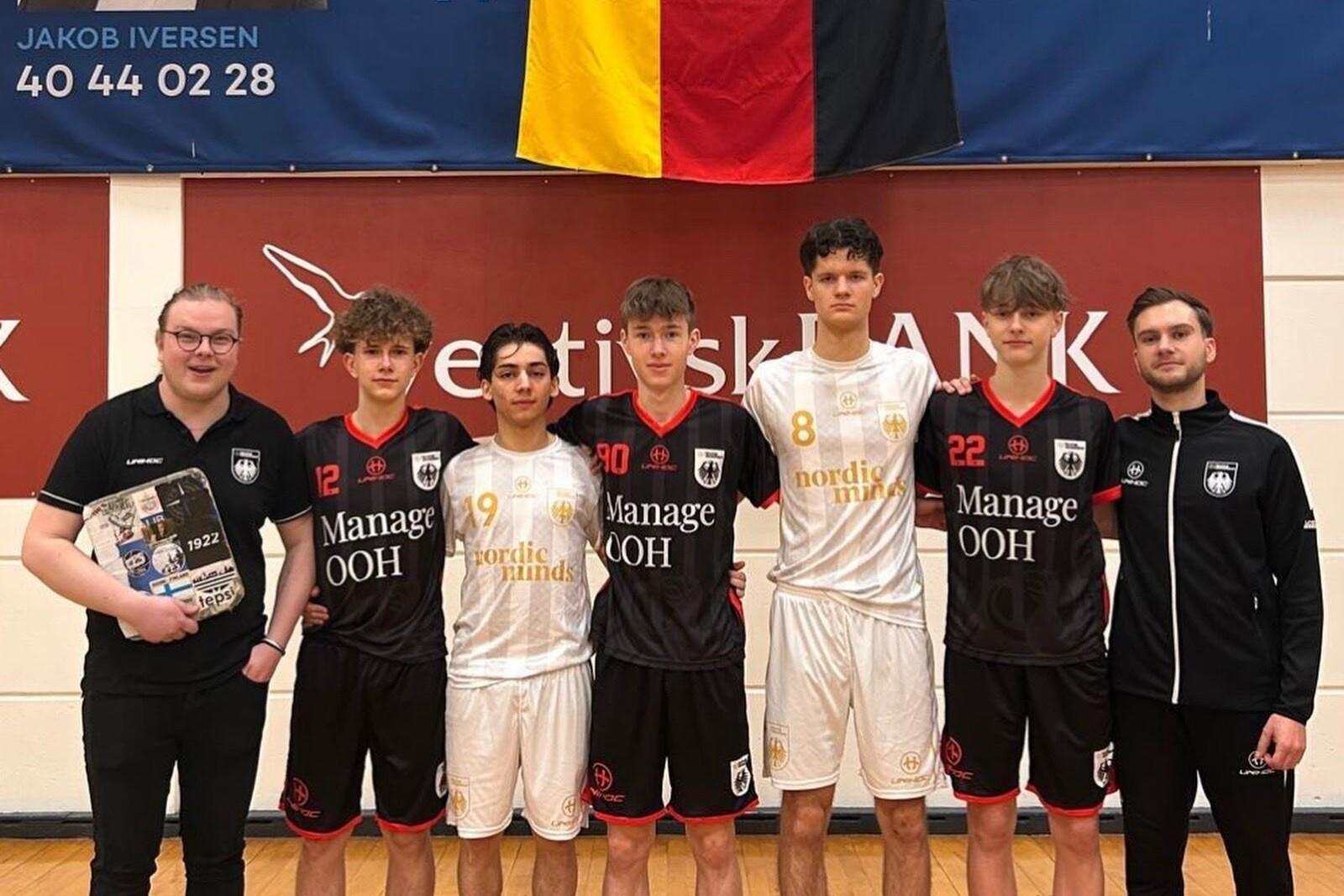 Sechs junge M&auml;nner in Sportoutfits stehen auf einer Halle, w&auml;hrend ein Banner mit der deutschen Flagge im Hintergrund h&auml;ngt.