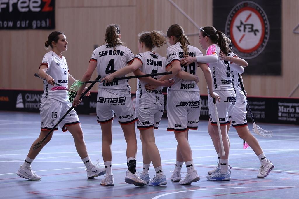 Sechs Frauen in Sportkleidung umarmen sich gl&uuml;cklich auf einem Hallenboden nach einem Spiel.