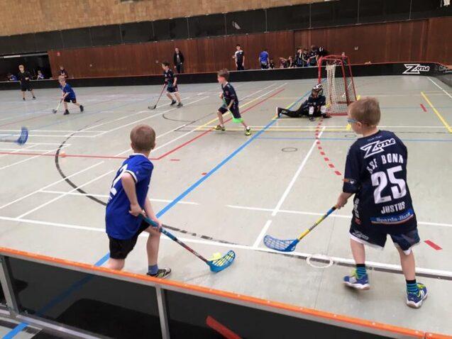 Sechs Kinder in Sportkleidung spielen Unihockey in einer Sporthalle; ein Torwart sitzt im Tor.