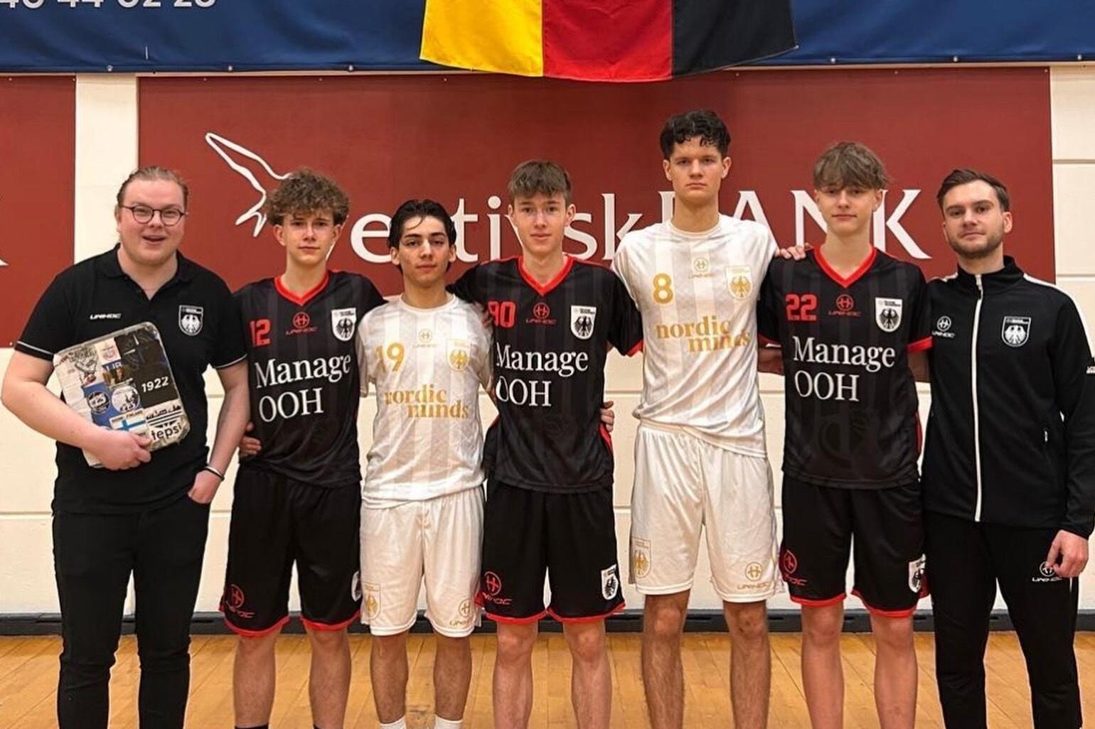 Sechs junge Männer in Sportoutfits stehen auf einer Halle, während ein Banner mit der deutschen Flagge im Hintergrund hängt.