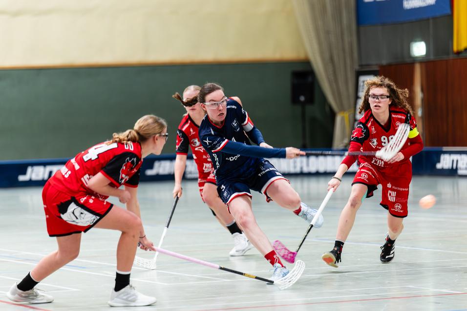 Vier Spielerinnen in Trikots k&auml;mpfen um einen Ball auf dem Boden eines Hallenbodens beim Unihockeyspiel.
