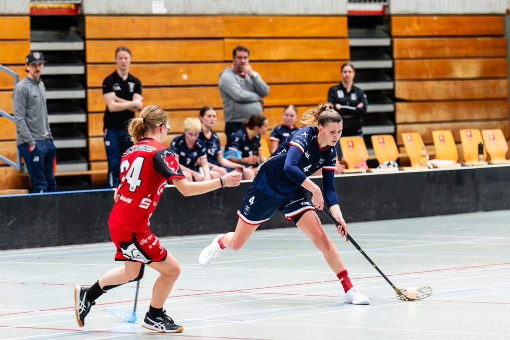 Zwei Frauen spielen in einem Sporthalle Unihockey; eine Spielerin dribbelt den Ball, die andere verteidigt.