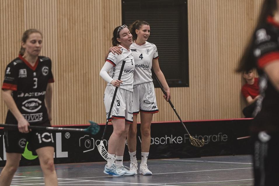 Zwei Spielerinnen im wei&szlig;en Trikot umarmen sich freudig auf einem Hallenboden w&auml;hrend eines Floorball-Spiels.