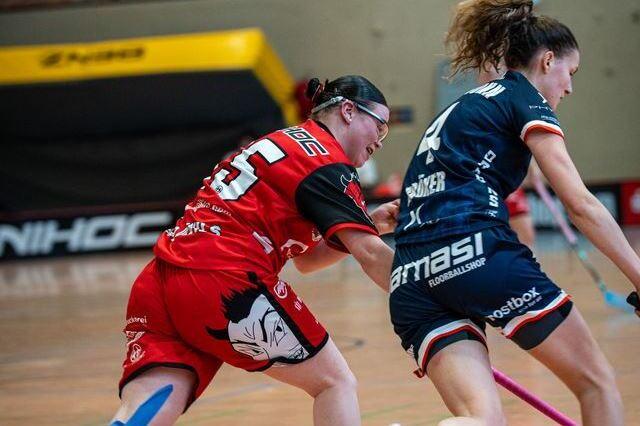 Zwei Frauen in Sportkleidung laufen in einem Hallenboden mit Schl&auml;gern in den H&auml;nden aufeinander zu.